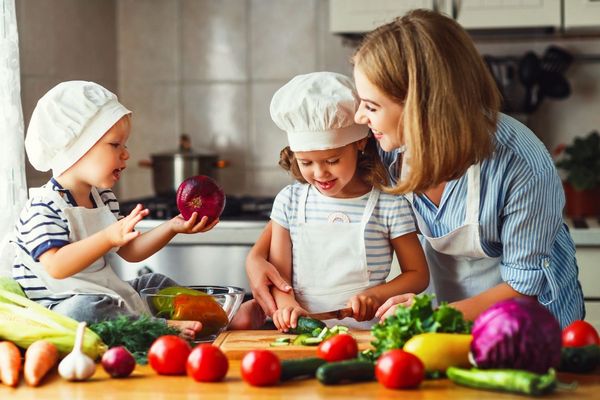 Egy konyhában anya kislányával és kisfiával zöldségeket tisztítanak, paradicsomokat, uborkákat, vörös káposztát, paprikát, sárgarépát, hagymát, salátát.