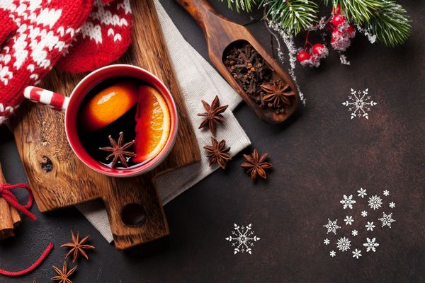 Egy szürke asztalon karácsonyi pulóver, karácsonyfa, mellette fakanálban csillagánizs, szegfűszeg, piros bögrében forralt bor naranccsal és csillagánizs.