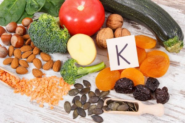 Egy asztalon K-vitamint tartalmazó élelmiszerek, cukkini, sárgabarack, aszalt szilva, tökmag, sárgaborsó, mandula, brokkoli, dió, paradicsom, mogyoró, spenót, paradicsom.