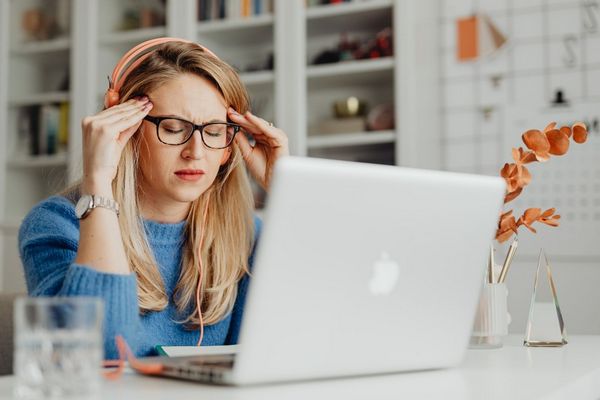 Egy fiatal nő az irodában a laptopján dolgozik, migrénes fájás miatt szemeit becsukja, fejét két kezével fogja.