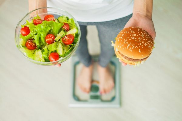 Egy férfi mérlegen állva egyik kezében egy tálban saláta, a másik kezében hamburger.