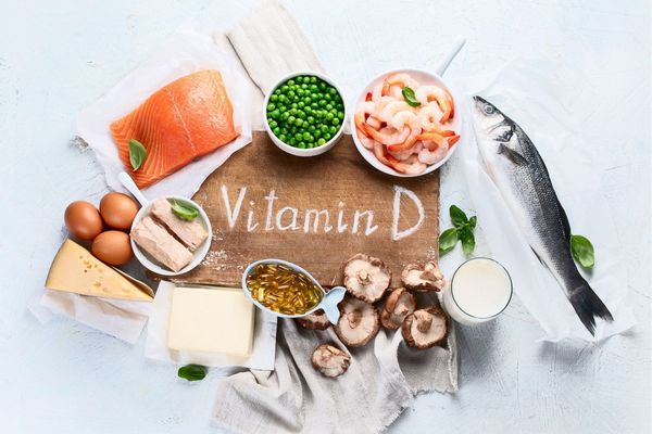 Egy asztalon D-vitamint tartalmazó élelmiszerek, lazac, tojások, sajt, borsó, rák, hal, spenót, tej pohárban, gomba, D-vitamin kapszulák.
