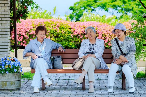 Egy virágos kertben három idős japán nő ülve beszélget és mosolyog.