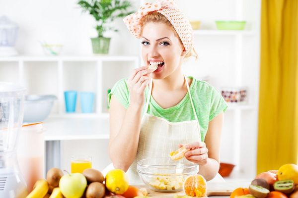 Egy fiatal nő a konyhában kendővel a fején narancsot eszik, az asztalon előtte sok gyümölcs, narancs, kivi, banán, citrom, alma.