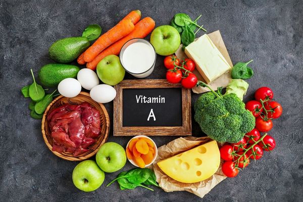 Egy szürke asztalon A-vitamint tartalmazó zöldségek és gyümölcsök, brokkoli, paradicsom, sajt, sárgarépa, avokádó, tojás, spenót, alma, alma, máj, aszalt sárgabarack.