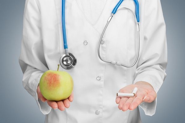 Egy fehér köpenyes orvos két kezét előre nyújtja, egyikben egy ketté rákot okozó tört cigaretta, a másikban egy alma.