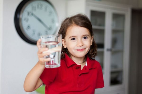 Egy kislány piros pólóban kezében egy pohár vízzel mosolyog.