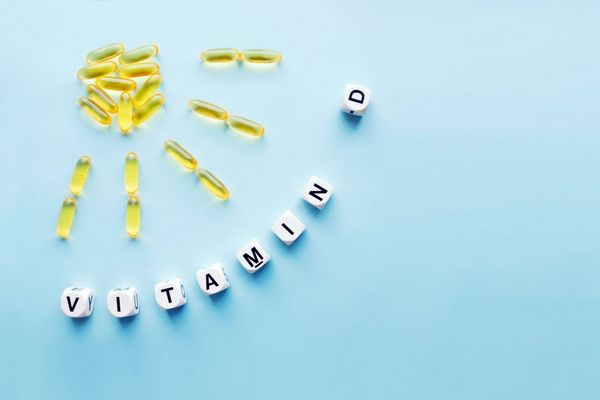 Egy kék háttér előtt D-vitamint tartalmazó kapszulák napot formáznak, előtte dobókockák D-vitamint felirattal.
