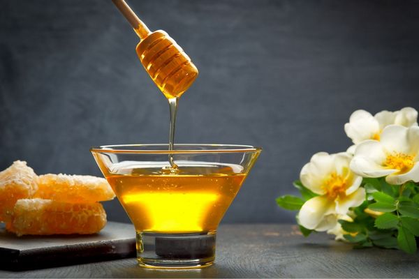 Egy asztalon egy üveg tálban méz, amiből egy mézcsurgatóval mézet vesznek ki, mellette virág és lépesméz.