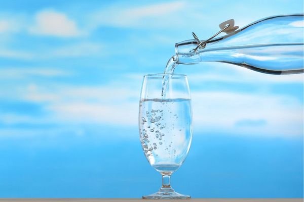 Egy felhős háttér előtt egy üvegből vizet öntenek egy üveg pohárba.