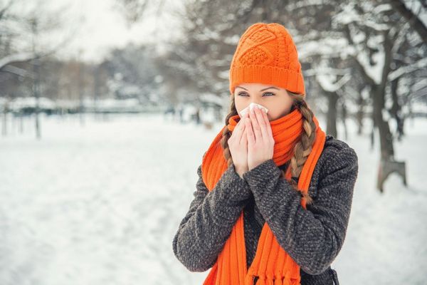 Egy téli havas parkban narancssárga sapkás és sálas lány az orrát papírzsepkendőbe fújja.