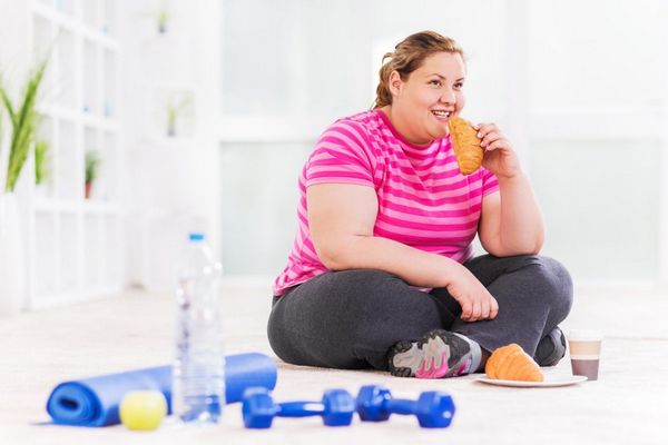 Egy túlsúlyos nő péksüteményt eszik, mellette fitneszmatrac és súlyzók.