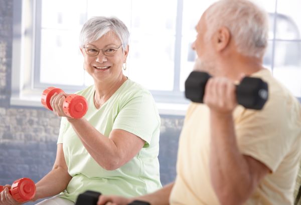 Idős nő és férfi ülve kézi súlyzókkal edzenek.