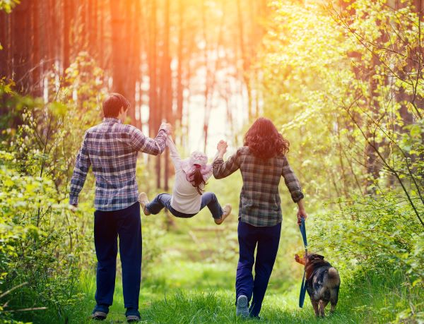 Erdőben sétáló család: szülők kockás ingben, közöttük egy kislány kézenfogva felemelve, kutya.