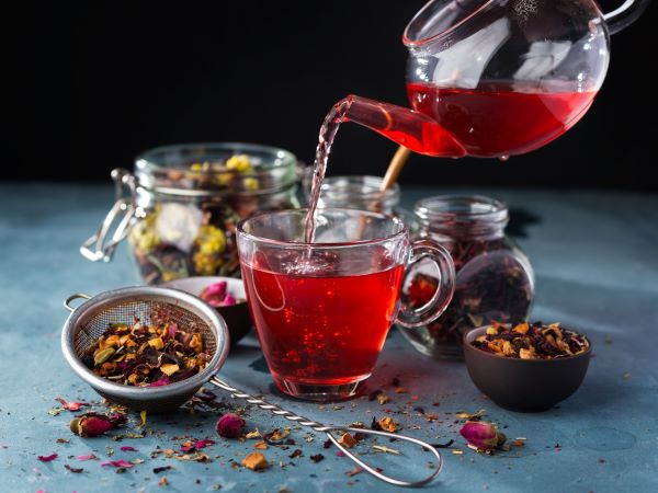 Üvegkancsóból üvegpohárba piros színű teát töltenek, körülötte üvegekben , tálkában, szűrőben és az asztalra szóródva színes teakeverékek.