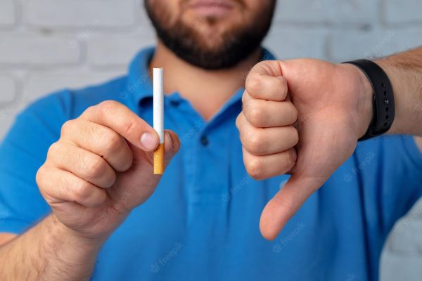 Férfi kék pólóban jobb kezében egy szál cigarettát tart, bal hüvelykujját lefelé fordítja.