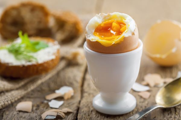 Faasztalon fehér tojástartóban megkezdett lágytojás, mellette kanál, tojáshéjak, háttérben barna kenyér.
