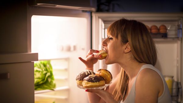 Fiatal nő este a nyitott hűtőajtónál titokban fánkot eszik.