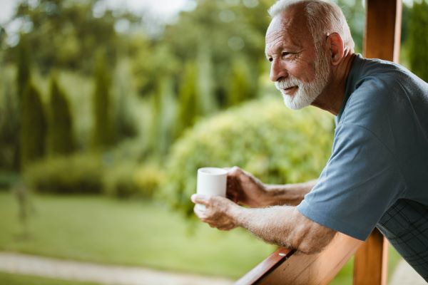 Idős, ősz szakállú férfi a teraszkorlátra támaszkodva kávécsészét tart és nézi a kertet.