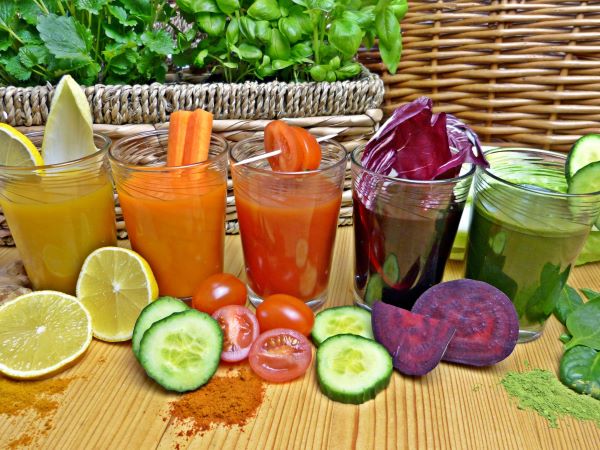 Asztalon 5 üvegpohárban különböző színű turmixitalok, tetejükön és előttük színes zöldségek, a háttérben egy hosszú fonott kaspóban zöld fűszernövények.