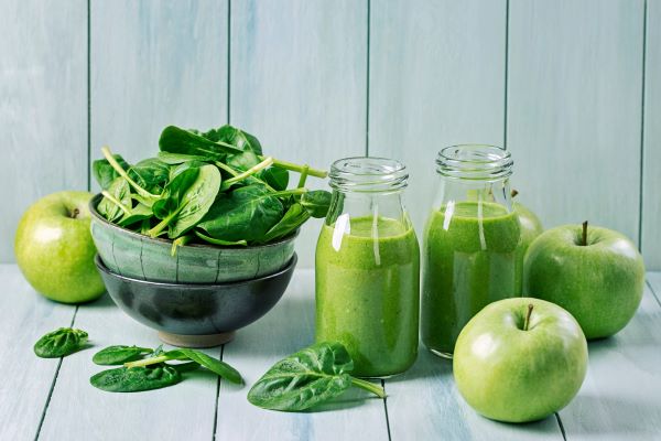 Konyhapulton egy tál spenótlevél, két üvegben zöld smoothie, mellette zöld almák.