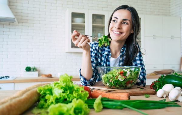 Fiatal nő vidáman eszik a konyhában salátát, körülötte a pulton zöldségek.