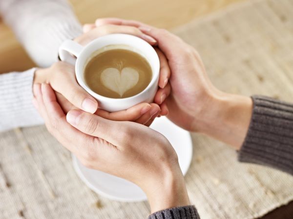 Női kezek fognak egy fehér kávéscsészét, a kávén szív mintájú tejszínnel, a női kezeket pedig férfikezek fogják közre.