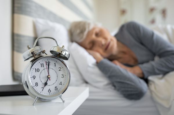 A háttérben egy idős nő alszik az ágyban, elöl az éjjeliszekrényen az ébresztőóra 7 órát mutat.