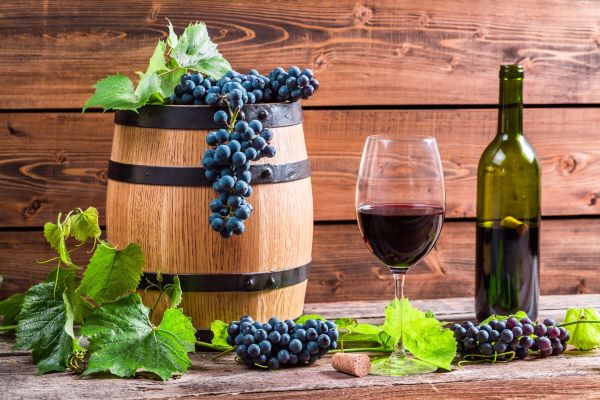 Kis fahordó tetején kékszőlő-fürtök, mellette egy üveg vörösbor, egy talpas pohárban vörösbor, kékszőlő-fürtök, szőlőlevelek, deszka háttér.