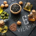 Fekete felületen krétával "E-vitamin" felirat, mellette napraforgómag, olajbogyó, lenmag, dió, kis üvegekben olajok.