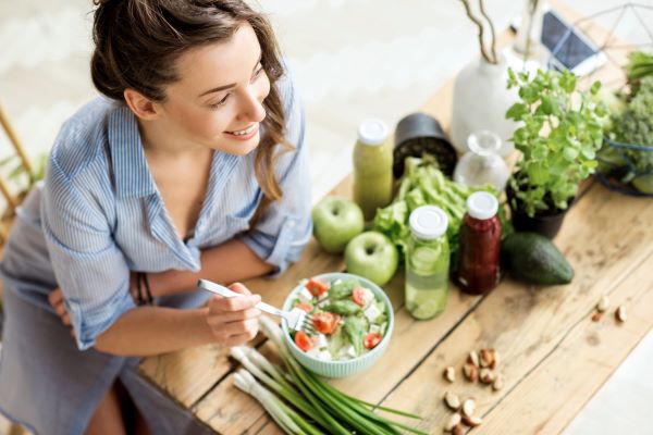 Mosolygó fiatal nő kék ingben ül az asztalnál és salátát eszik tálból, mellette zöldhagyma, zöld alma, kesudió, cserepes fűszernövények, üvegekben salátaöntetek.