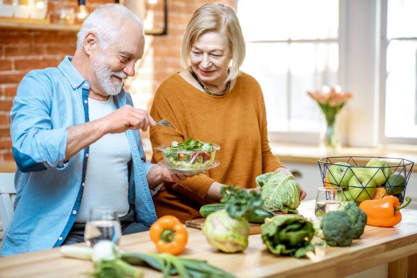 Idős házaspár a konyhapultnál zöldségekkel körülvéve salátát készít.