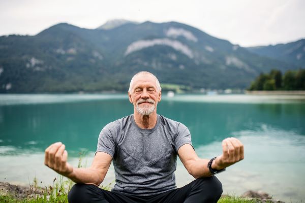 Idős férfi törökülésben meditál, mögötte tó és hegyek.