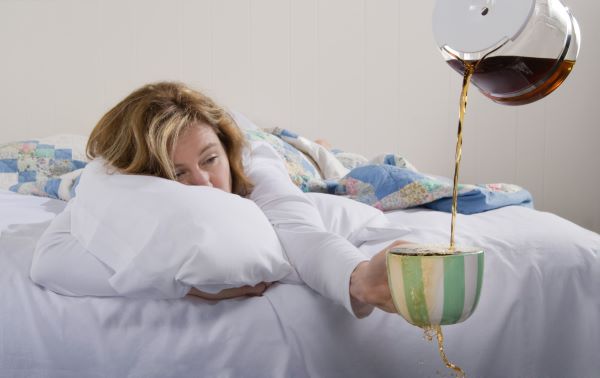 Álmos nő hason fekszik az ágyban és egyik kezét kinyújtva a paplan alól tart egy csíkos bögrét, amelybe valaki kávét tölt, ami túlcsordul.
