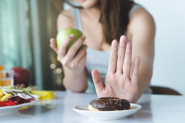 Fiatal nő az asztalnál ülve egyik kezében zöld almát tart, másik kezével eltol magától egy tényért, melyben csokis fánk van.