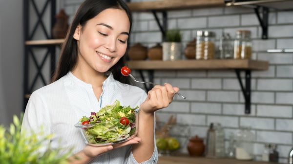 Fiatal nő a konyhában egy tál salátát tart mosolyogva, másik kezében egy villa végén koktélparadicsom.