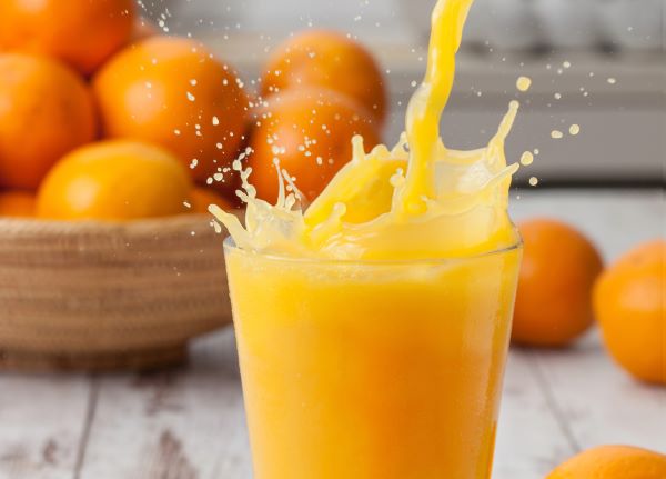 Egy pohárból kifröccsenő narancslé, háttérben egy tál teli naranccsal.