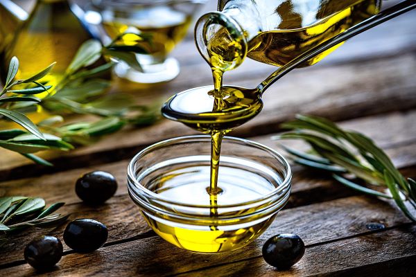Valaki üvegből olívaolajat önt egy kanálba, amiből az olaj egy kis üvegtálba csorog, amely rusztikus faasztalon áll, körülötte néhány olajbogyó és olajfaág.