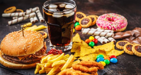 Asztalon különféle egészségtelen ételek: hamburger, sült krumpli, fánk, chips, kóla, színes cukorkák, sütemények.