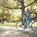 Idősebb nő kerékpározik egy parkon keresztől, fák között.