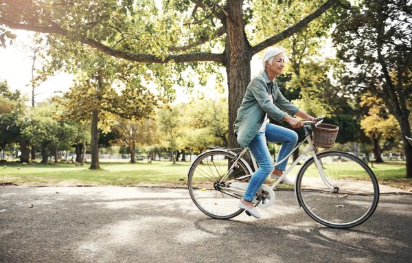 Idősebb nő kerékpározik egy parkon keresztől, fák között.