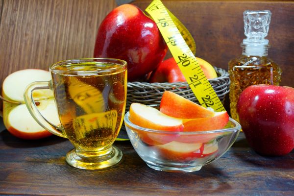 Egy pohár és egy üveg tele almaecettel, egy kis kosárban piros almák, rajtuk mérőszalag, mellette egy egész és egy fél alma, kis üvegtálban almaszeletek.