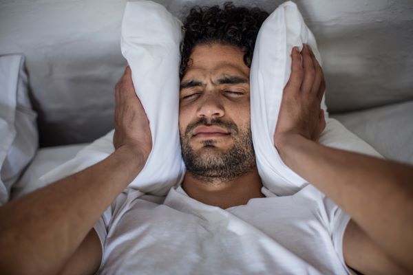 Szakállas férfi ágyban fekve kétoldalt a füléhez szorítja a párnát.