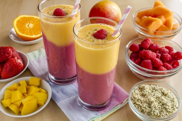 Két magas pohárban kétszínű turmix: alul rózsaszínű, felül sárga, benne szívószál, tetején 1-1 szem málna, körülötte kis tálkákban a hozzávalók: mangó, málna, narancs, eper.