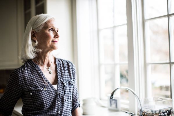 Idősebb ősz hajú nő kockás ingben áll a konyhában az ablaknál és néz kifelé.