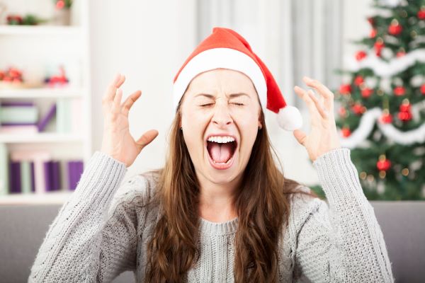 Szobában a kanapén kötött pulóverben és mikulássapkában ülő nő felemelt karokkal és csukott szemmel kiabál, háttérben karácsonyfa, könyvespolc.