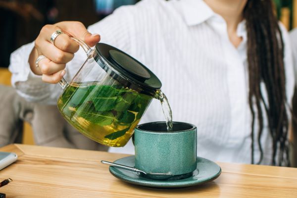 Hosszú hajú nő asztalnál ülve üvegkancsóból zöld teát tölt egy csészébe.