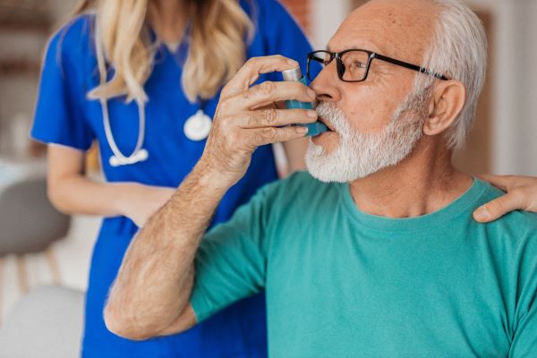 Idősebb szakállas férfi inhalálóval lélegzik, mellette fiatal kék köpenyes doktornő áll sztetoszkóppal a nyakában.