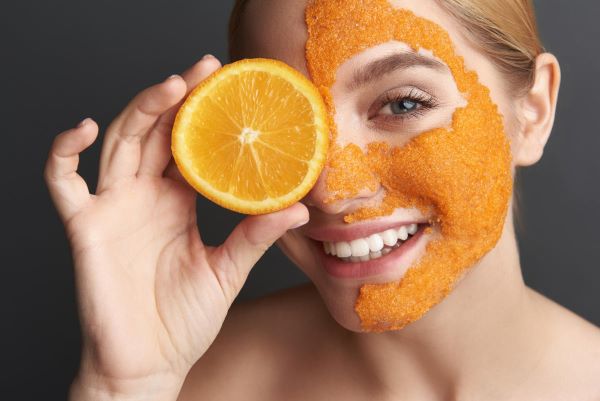 Fiatal nő mosolyogva az egyik szeme elé tart egy fél narancsot, miközben az arca másik felét narancshéjas pakolás borítja,