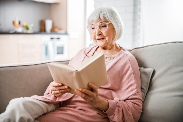 Idős szemüveges nő ül egy kanapén kezében egy nyitott könyvvel és olvas.